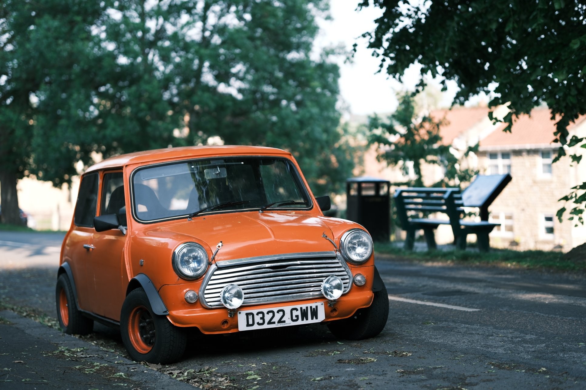 A old, small, orange car (Mini) is parked on the side of the road, its wheels turned as if to drive off. The bright orange of the car contrasts with the muted greens of the surrounding trees and the gray of the road.  A old, small, orange car (Mini) is parked on the side of the road, its wheels turned as if to drive off. The bright orange of the car contrasts with the muted greens of the surrounding trees and the gray of the road.